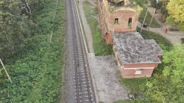 Golden hour flyover of overgrown two-storey railway signal building and office block beside single rail in Bangladesh countryside