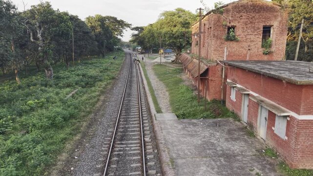 Elevated daytime view along overgrown rail beside old red service block with pedestrians in rural India