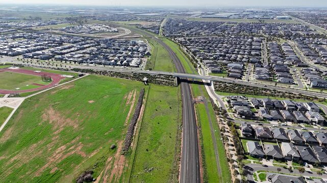 Aerial high-angle panorama showing regional railway tracks and an overpass bridge cutting through modern master-planned housing estates and sports grounds in Truganina, Melbourne, Australia.