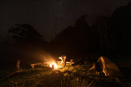 A young man bends over to place wood on a campfire with an tent to the side and stars above