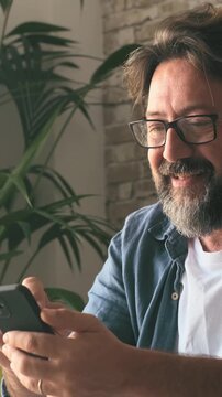 Cheerful middle-aged man with glasses enjoying online content, laughing and texting on his mobile phone at home. Modern lifestyle and connection technology for adult people
