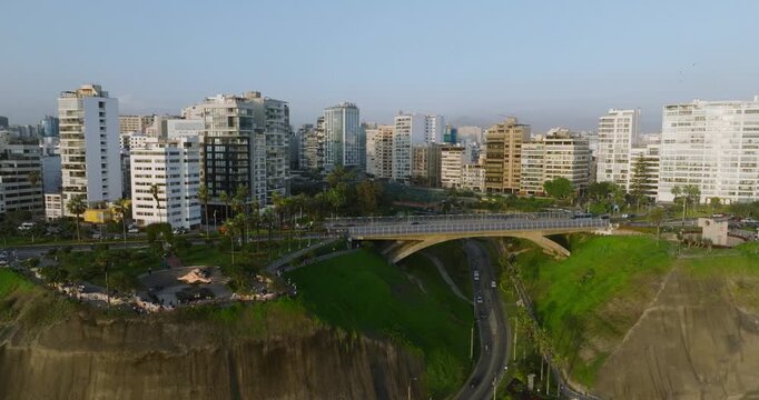 Aerial view of the coastal cliffs and modern skyline of Miraflores district in Lima, featuring a bridge over the highway and lush green parks in Lima, Callao Region, Peru.