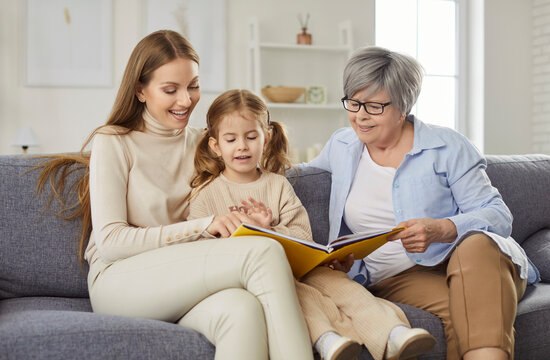 Three generations of women sitting together on sofa at home and reading book. Smiling grandmother, mother and little girl enjoying family time and learning together. Family leisure and storytelling.