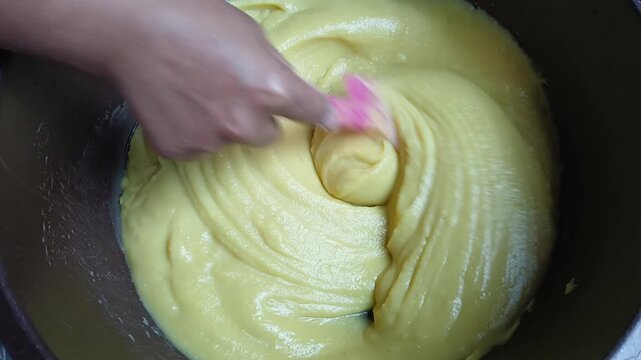 Hand stirring smooth yellow cake batter or custard cream in a bowl using a pink spatula