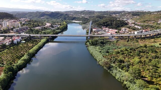 Aerial view of the Ponte Rainha Santa Isabel spanning the Mondego River, surrounded by lush green banks and urban development in Coimbra, Coimbra, Portugal.