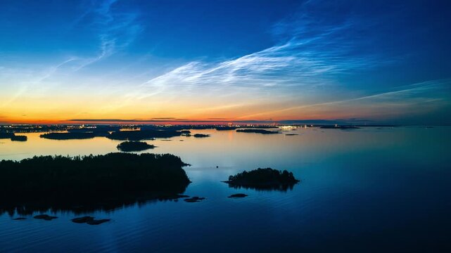 Aerial view of the Espoo archipelago featuring calm waters, small forested islands, and vibrant noctilucent clouds during a summer twilight in Espoo, Uusimaa, Finland. Hyperlapse.
