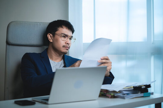Businessman reviewing documents while working on a laptop in a modern office, showing concern and stress while analyzing business information, financial problems, workload, deadlines