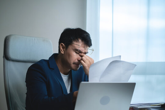 Businessman reviewing documents while working on a laptop in a modern office, showing concern and stress while analyzing business information, financial problems, decisions. 