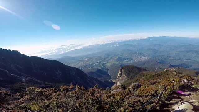 Wide panning shot looking down from the rugged trail and alpine scrub of Mount Kinabalu over the expansive Kundasang valley and distant ranges under bright sunlight.