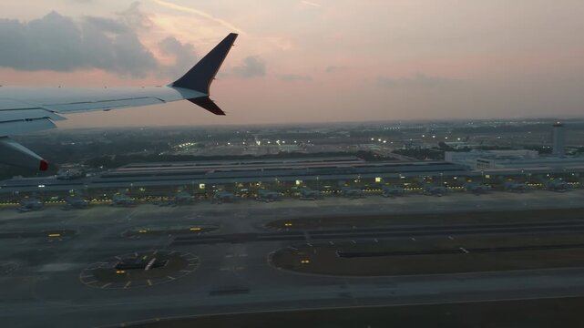 Passenger window view showing an aircraft wing passing over runways, taxiways and a terminal complex beneath a warm evening sky.