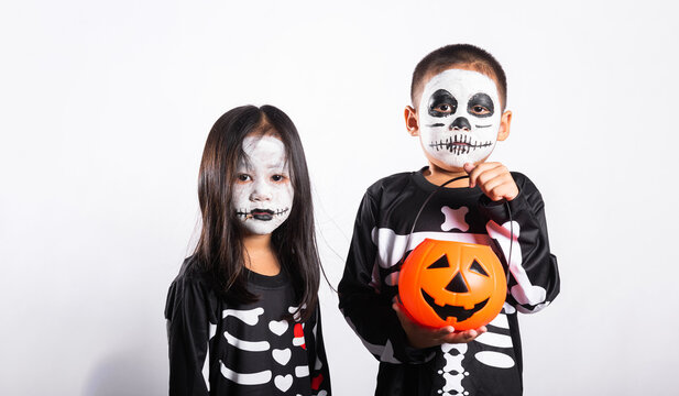 Halloween kid celebration involving young boy and girl wearing skeleton costumes and face paint while holding pumpkin bucket against plain white background for autumn holiday party event