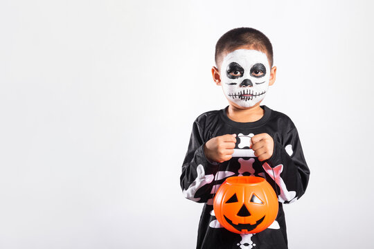Halloween festival celebration featuring happy kid wearing skeleton costume and painted face holding orange pumpkin bucket while standing isolated against plain white background illumination