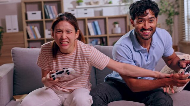 Asian couple playing video game with controller in living room on sofa, playful competition, smiling man and distressed woman showing excitement and animated expressions during home gaming session