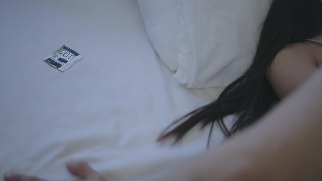 Hand Reaching Across White Bed Toward Condom Packet, Safe Sex and Sexual Health Bedroom Relationship Care, Sleeping Woman Facing Financial Stress, White Shirt, Banknote, Dark Hair, Privacy