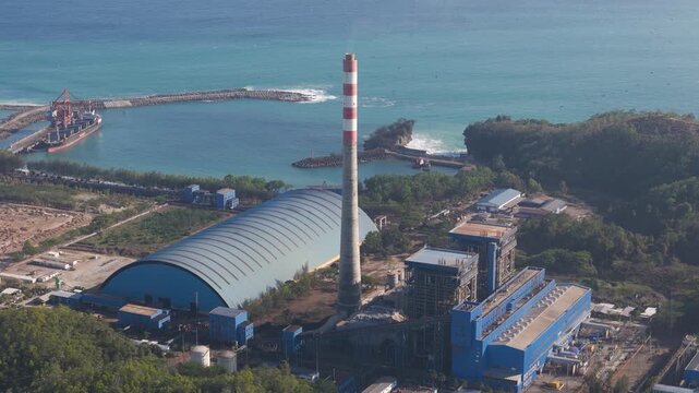 Aerial view of a coastal power plant featuring a tall striped chimney, a large arched storage facility, and industrial buildings situated along the coastline in Pacitan, East Java, Indonesia.