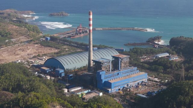 Aerial view of the Pacitan Power Plant featuring a tall chimney and blue industrial buildings situated along the coastline in Pacitan, East Java, Indonesia.