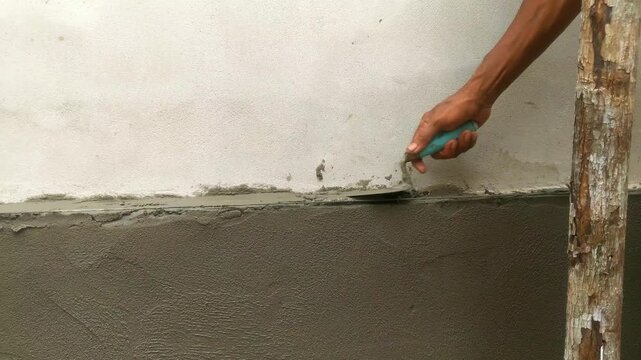 A construction worker applying wet cement plaster to a wall using a metal trowel.