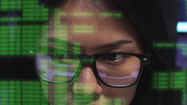 Close up of a female programmer in glasses reading moving lines of green code that scroll across her face, working late at night in a dark office.