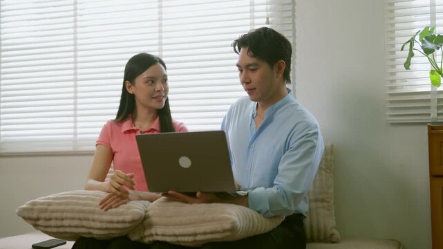 Young asian couple using a laptop computer for online shopping and planning their vacation together in a modern living room