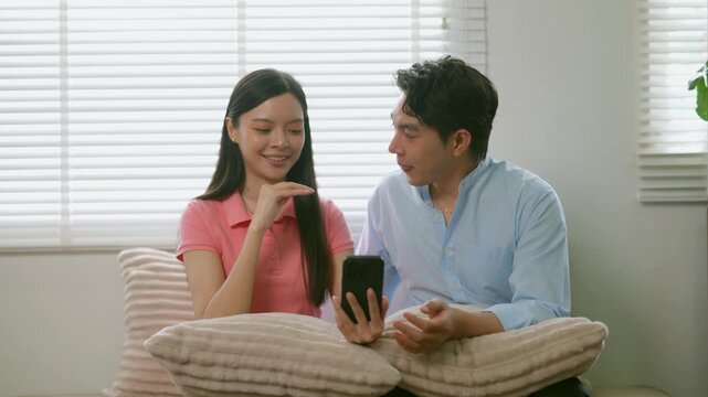 Young asian couple enjoying their time together at home, happily browsing social media and shopping online on a modern smartphone