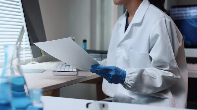 Female scientist in a lab coat and gloves analyzing research documents at a modern laboratory desk with a computer
