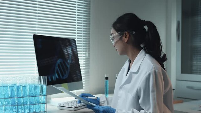 Asian scientist analyzing DNA helix on computer and holding test tube with blue liquid in a modern laboratory research facility
