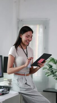 Smiling asian businesswoman using a digital tablet leaning on her desk in a modern corporate office and checking company data