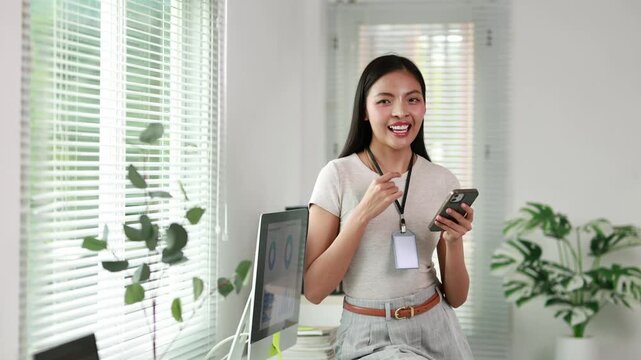 Young Asian businesswoman using a smartphone and dancing joyfully to music in a modern bright office at her desk