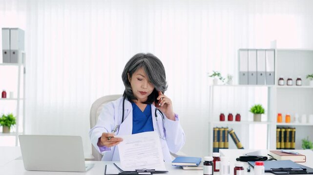 asian female doctor in scrubs with a stethoscope reads a medical report on a clipboard while talking on the phone in a hospital  office.
