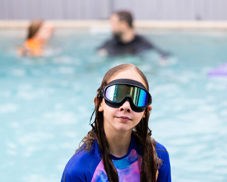 Young girl standing in a swimming pool wearing a rash guard and large reflective goggles