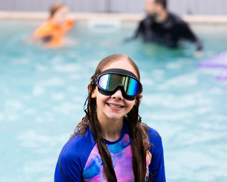 Young girl standing in a swimming pool wearing a rash guard and large reflective goggles