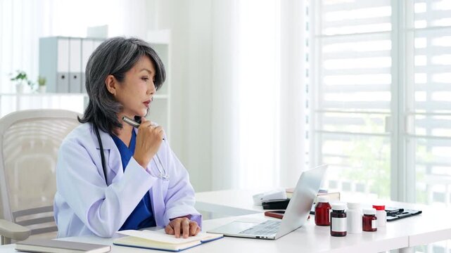 Doctor working in medical office using laptop computer and writing or taking note in notebook.