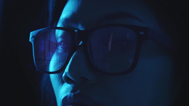 Close-up image of a female cybersecurity analyst in a dark room at night. Code and digital data are reflected in the lenses of her black-rimmed glasses as she stares at a screen.