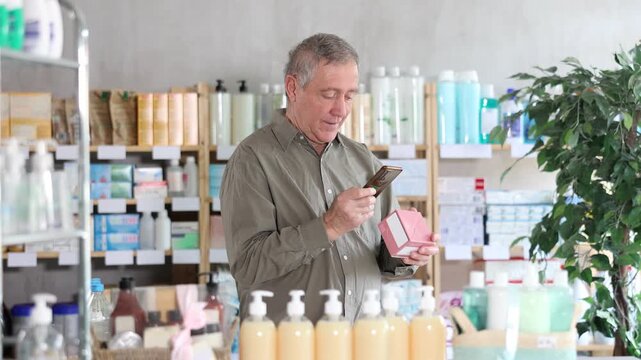 Focused elderly man in pharmacy scanning barcode on box of pills with smartphone, carefully checking dosage, side effects, or contraindications before purchase, ensuring safe self-medication 