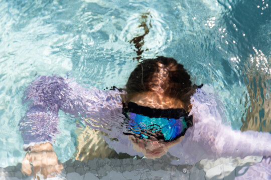 Young girl wearing reflective blue-green goggles submerged underwater in a clear blue pool