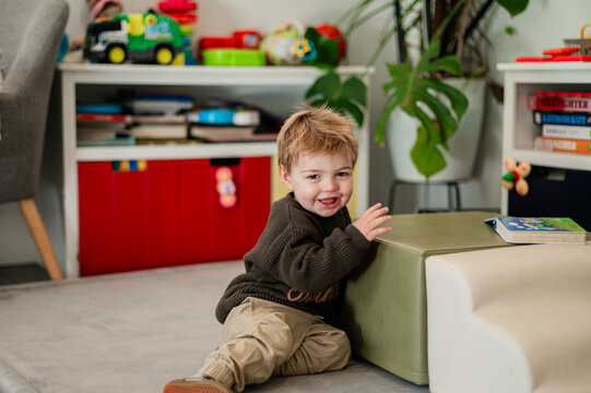 A young boy sits on the floor, smiling while playing with a green cube in a room with toys