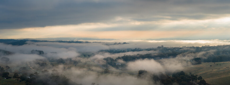 Morning fog drifting across rolling countryside beneath dramatic overcast sunrise skies