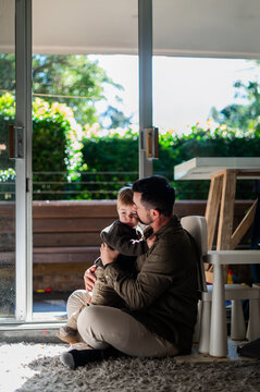 A father holds his young son on the floor, sharing a gentle moment in a bright room