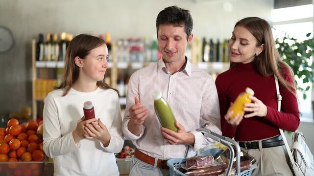 In healthy eating store, positive family of customers chooses smoothie and actively argues about purchase. Mom, dad and daughter view fruit and herbal mix cocktail, read expiration date of product