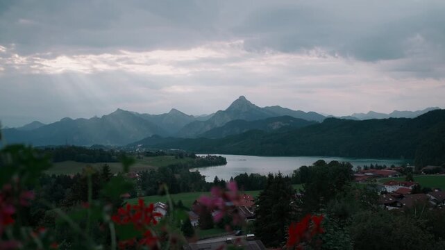 Slow dolly move on a balcony near F&uuml;ssen, red geraniums in the foreground giving way to Lake Wei&szlig;ensee and the Bavarian Alps in bright, soft daylight. A welcoming Allg&auml;u view from the holiday balcony,