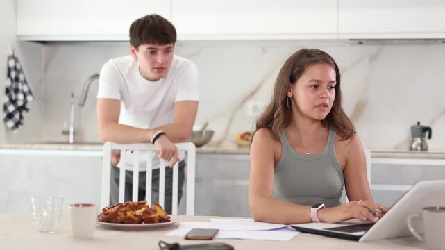 Sad young girl working on laptop while her boyfriend is quarreling with her standing behind in the kitchen