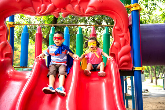 Two children in superhero masks sliding down a colorful playground slide.