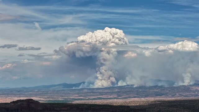 4K drone timelapse capturing an aggressive pyrocumulus cloud forming from a raging wildfire. The massive, mushroom-like smoke plume towers high into a blue sky above a vast, dry mountain range.