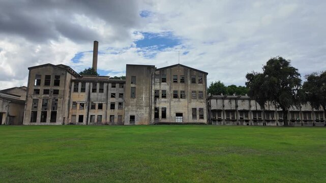 Wide angle footage shows the empty, deteriorating shell of an abandoned paper mill in Kanchanaburi, Thailand. Overcast sky, green grass, and ominous lighting set a moody, industrial scene.