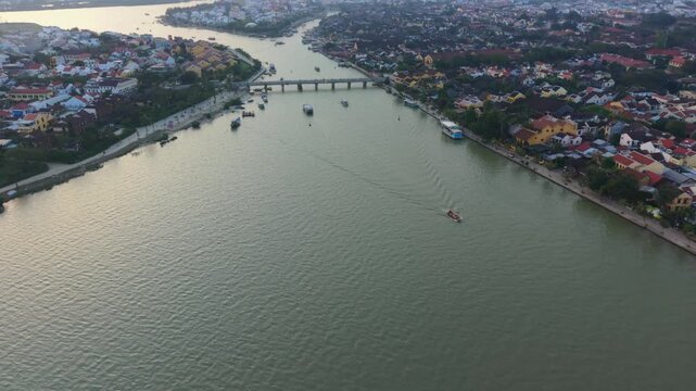 Vertical screen Aerial View Hoi An River With Boats Crossing Toward Bridge, Boat Wakes On Green Water, Scattered Vessels Moored Along Banks, Evening Light Softening Scene, Maritime Movement