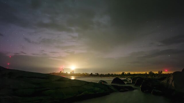 Perseids and satellites cross the night sky as a distant thunderstorm lights the horizon behind Ut&ouml; Lighthouse. The Baltic Sea rolls below as darkness fades into sunrise.
