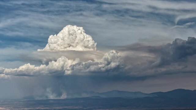 4K drone timelapse capturing a dramatic pyrocumulus cloud forming over a distant mountain range. A towering white convective column bursts upward through a thick, dark layer of wildfire smoke.