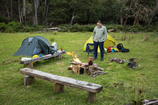 A young man in his twenties stands in front of a small campfire with tents and forest behind