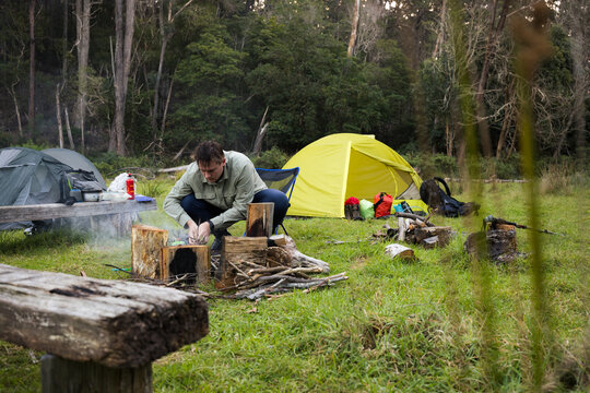 A young man in his twenties crouches down to add sticks for starting a campfire