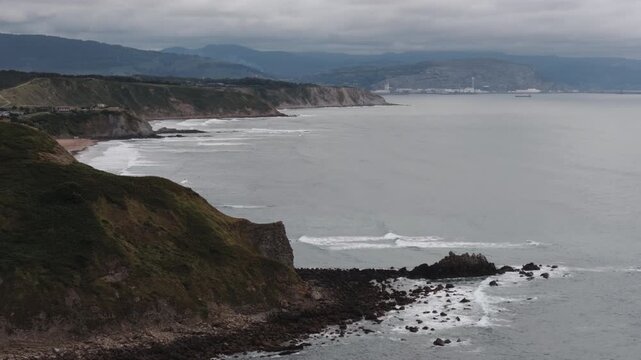 Coastal landscape with rolling hills, a sandy beach, and waves breaking on a rocky shore under an overcast sky